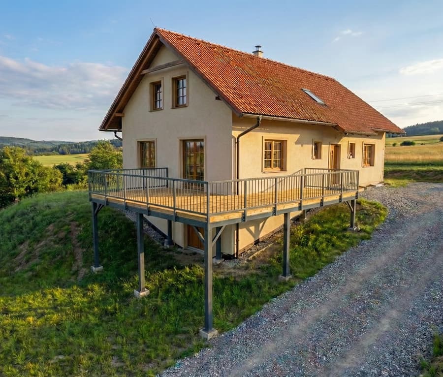 Ocelová terasa na míru od KOVOSET – Kvalitní konstrukce Pohled na vyvýšenou ocelovou konstrukci terasy u rodinného domu ve svahu se zábradlím a zinkovaným povrchem.