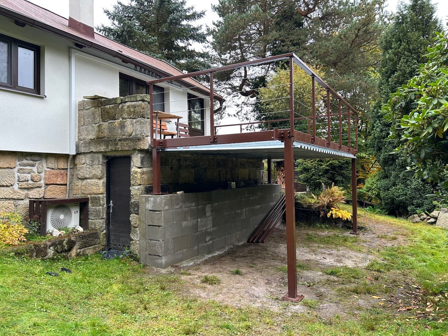 Od těžkých ocelových konstrukcí až k vaší terase Vyvýšená ocelová terasa na sloupech s hnědým nátěrem a lankovým zábradlím u rodinného domu.